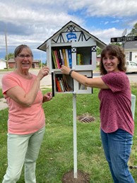 2 people pose in front of a Little Library